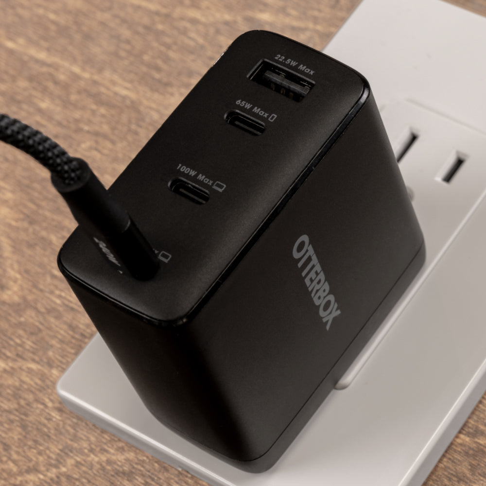 A black Otterbox 4-port USB-C fast charger plugged into a white wall outlet on a wooden surface. The charger features two USB-C ports, one 140W Max and one 65W Max, with the Otterbox brand name printed in white on the side.