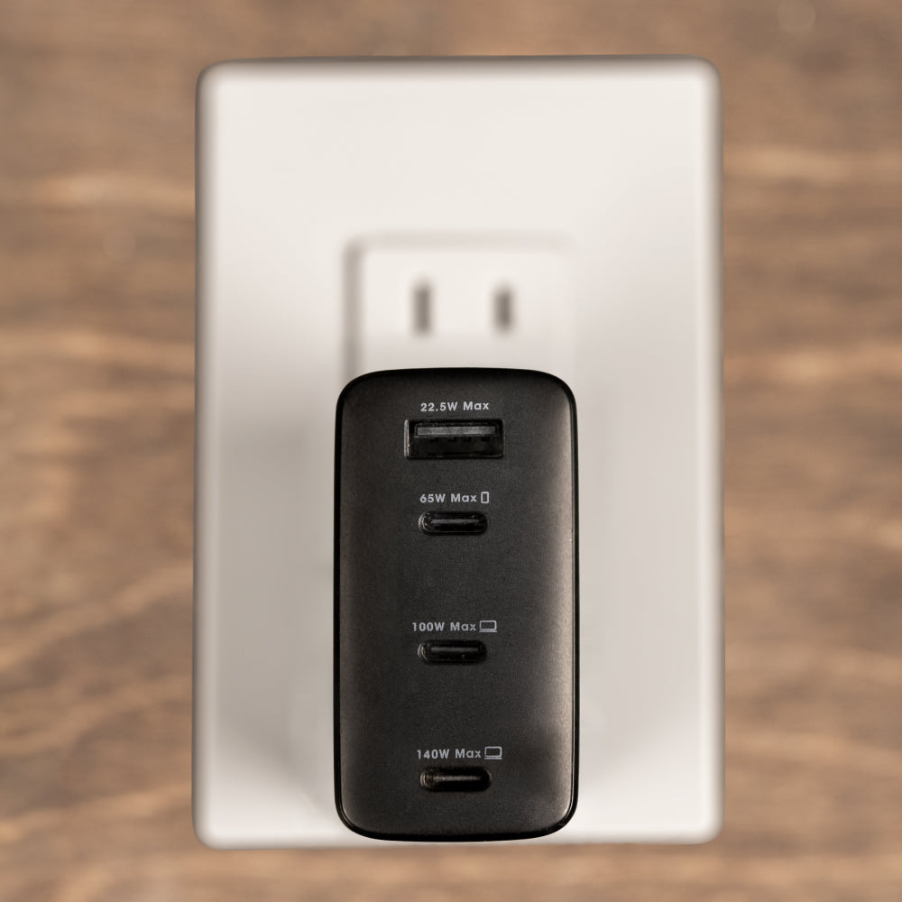A black multi-port USB charger plugged into a white wall outlet on a wooden surface. The charger has four ports labeled 22.
