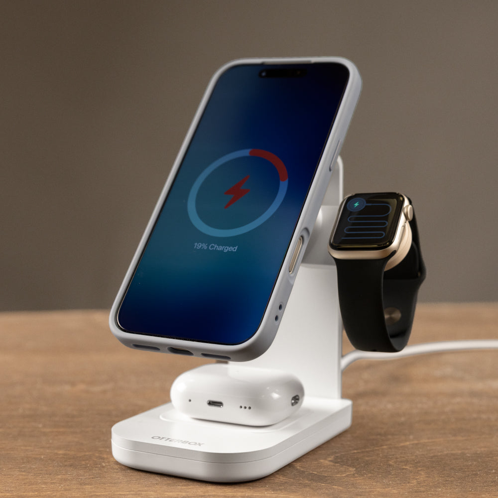 A white wireless charging stand on a wooden surface, charging a smartphone, AirPods, and a smartwatch. The smartphone screen displays a blue charging interface, and the background is a soft, warm gray-brown gradient.