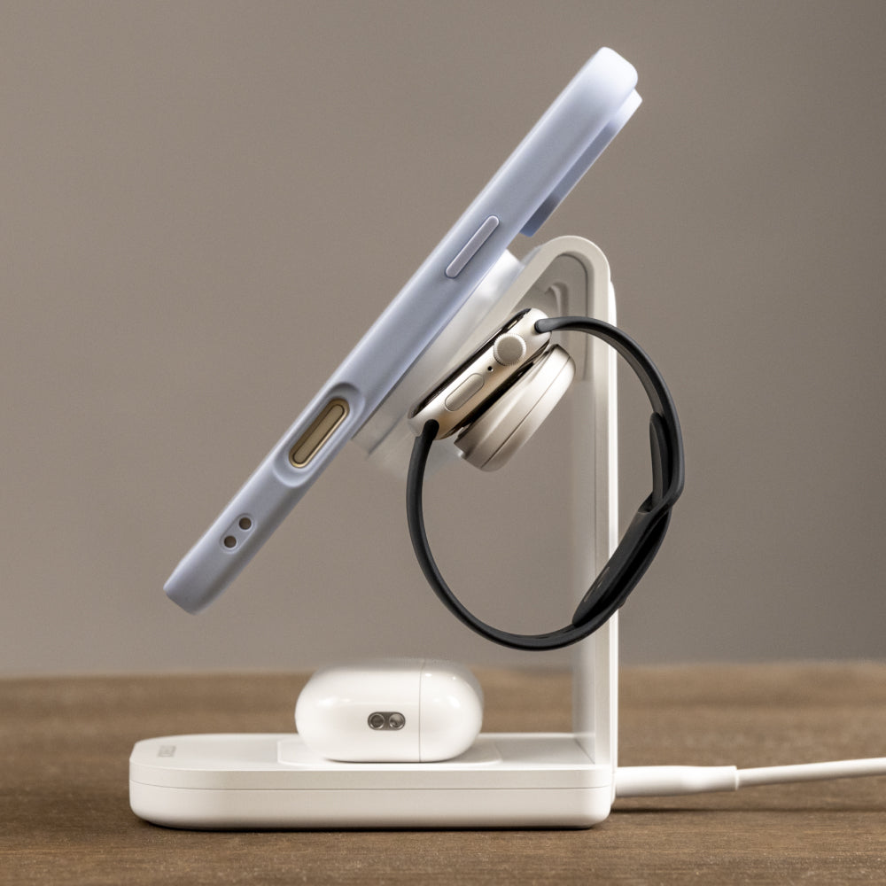 A white magnetic wireless charging stand on a wooden surface, shown charging a light blue smartphone, black over-ear headphones, and white earbuds in a clean studio setup.