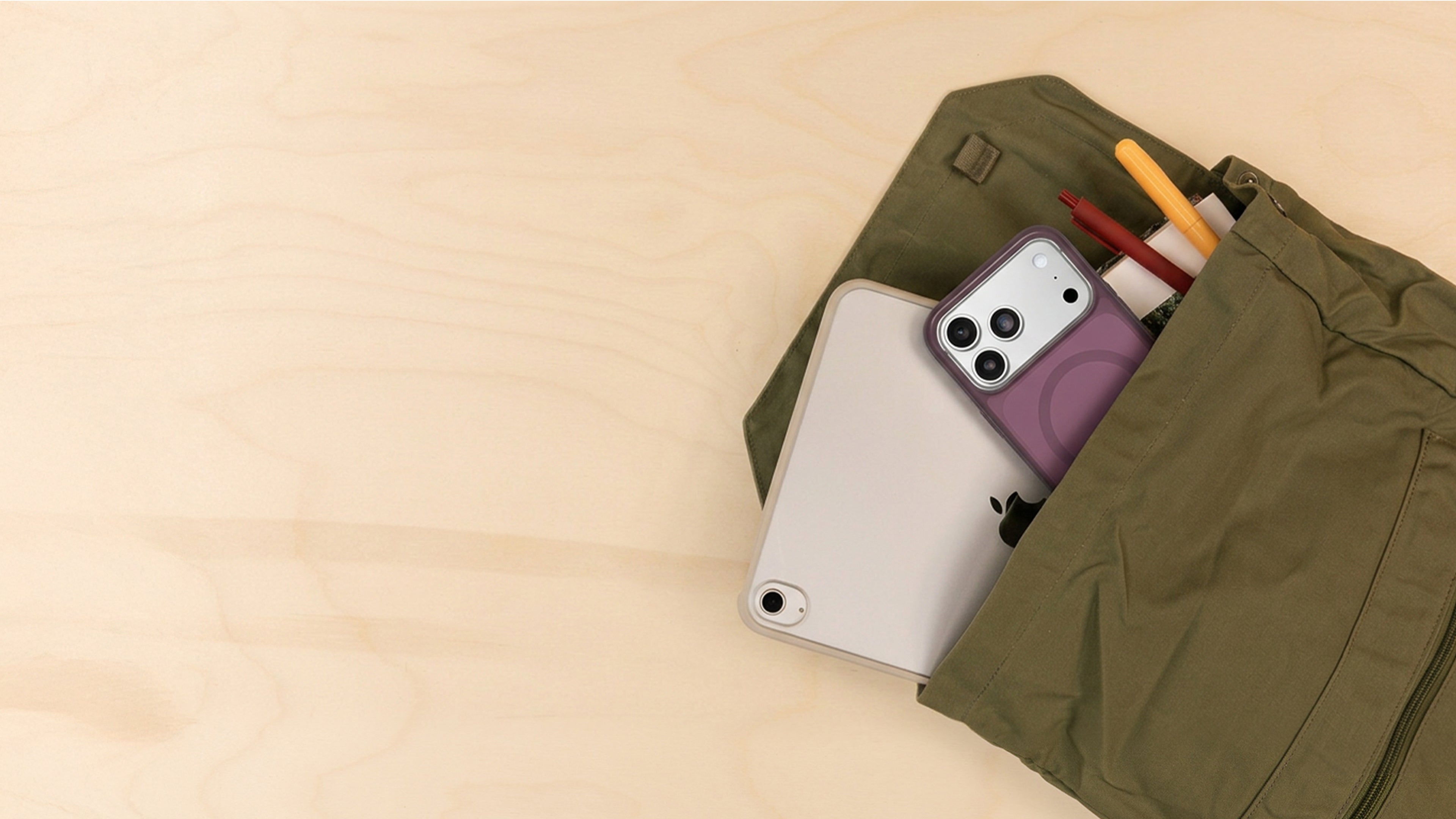 An olive green fabric pouch or organizer laid on a light wood surface, partially open to reveal a white tablet, a purple phone case with a camera cutout, and yellow and red pens.