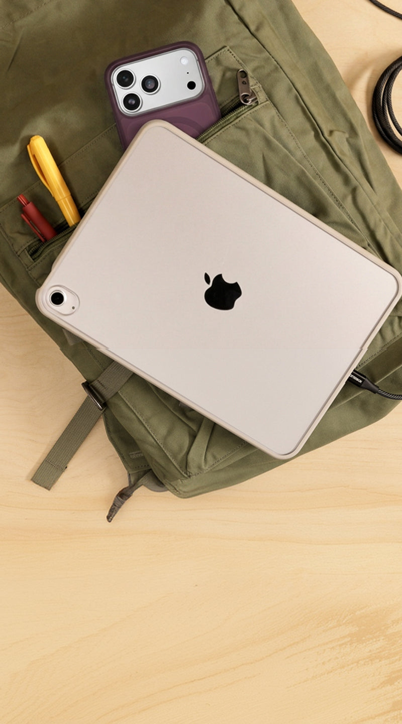 A silver Apple iPad with a black Apple logo lies on top of an olive green backpack, which is placed on a light wood desk. A purple smartphone is tucked into the backpack's side pocket, along with a yellow pen and other small accessories.
