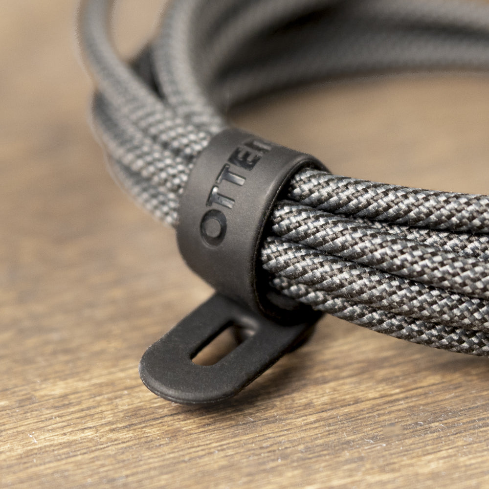 Close-up of a braided gray and black cable secured with a matte black plastic Otter loop, resting on a wooden surface.