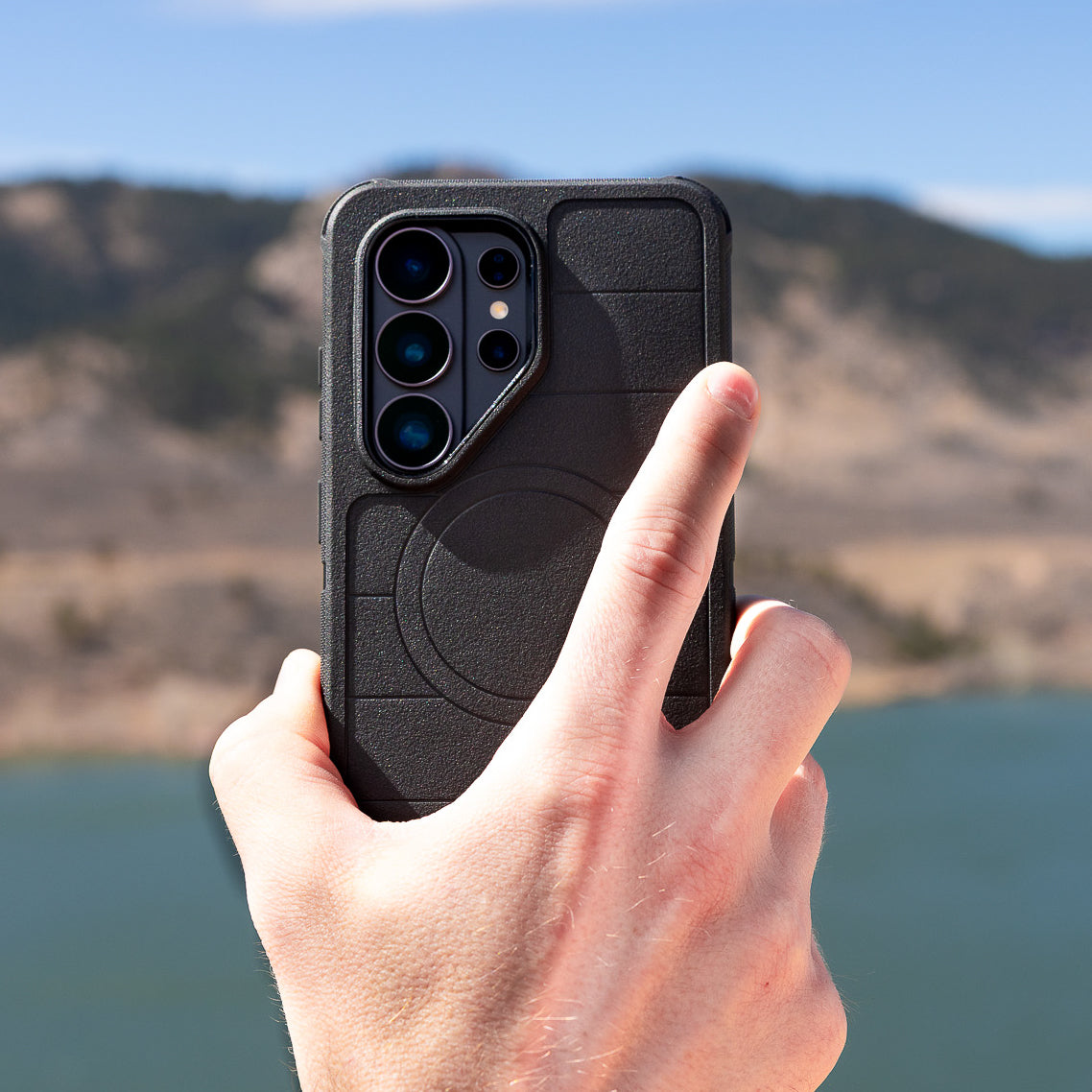Hand holding a black phone case with a scenic background of mountains and water.