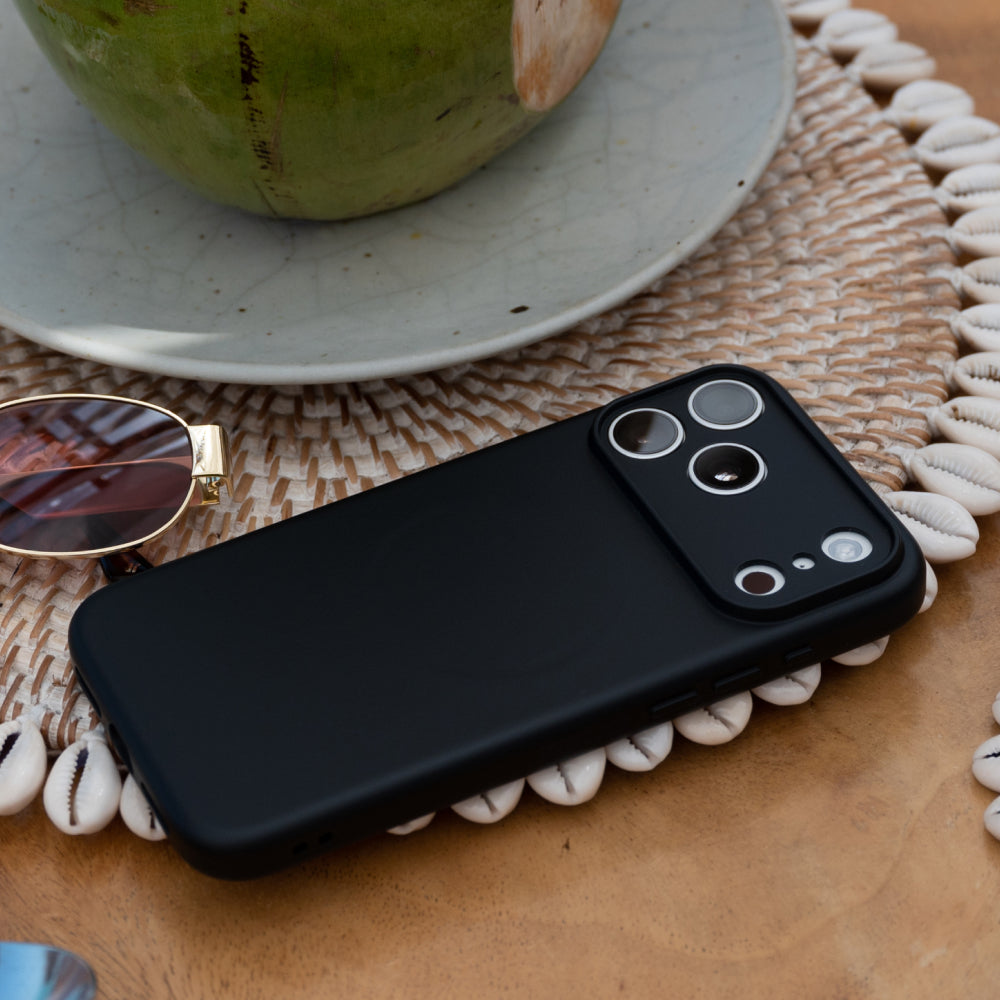 A matte black phone case on a smartphone, styled on a woven placemat with a coconut, sunglasses, and a ceramic plate in the background.