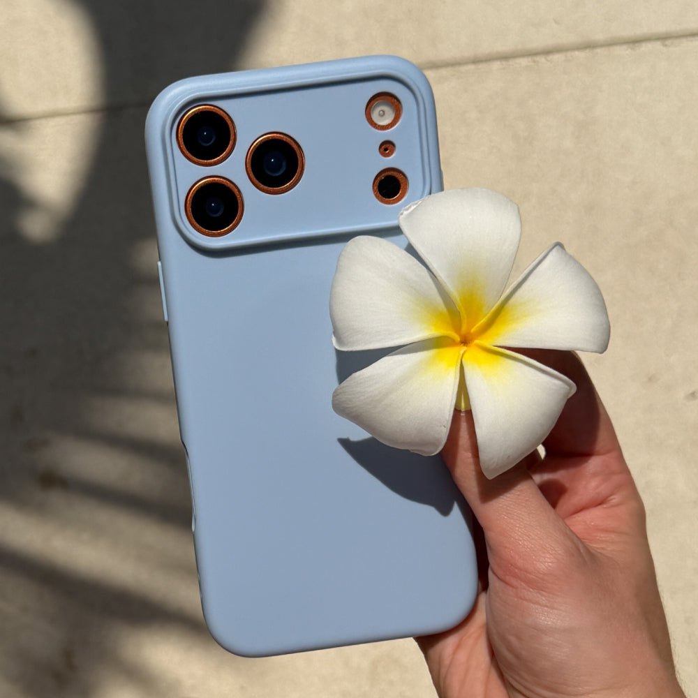 A light blue phone case with a raised camera cutout, shown being held by a hand against a sunlit beige stone background. A white plumeria flower is positioned in front of the phone case.