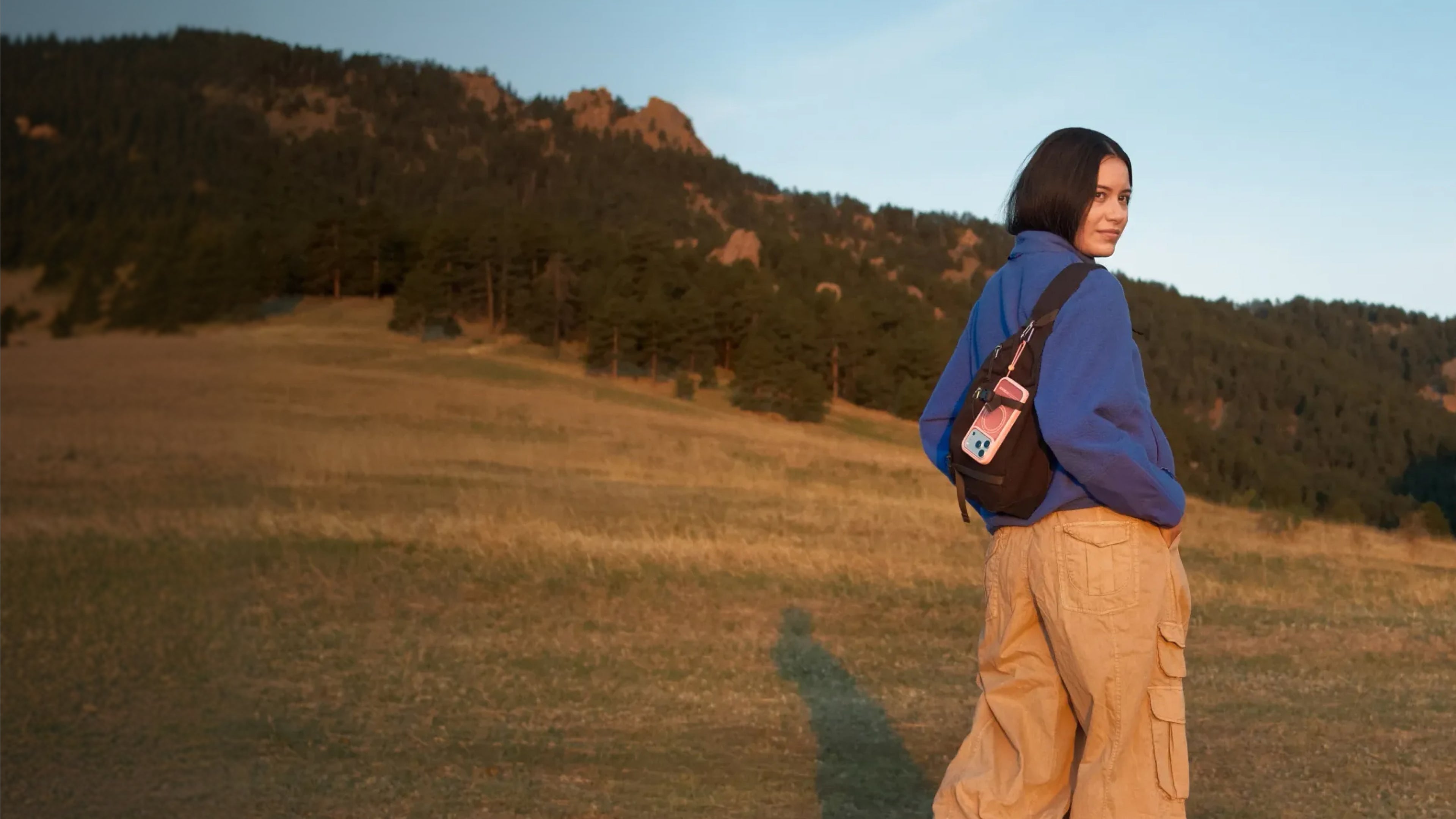 A woman stands in a grassy field with mountains in the background, wearing a blue hoodie, beige cargo pants, and a brown shoulder bag with a Sole series case attached.