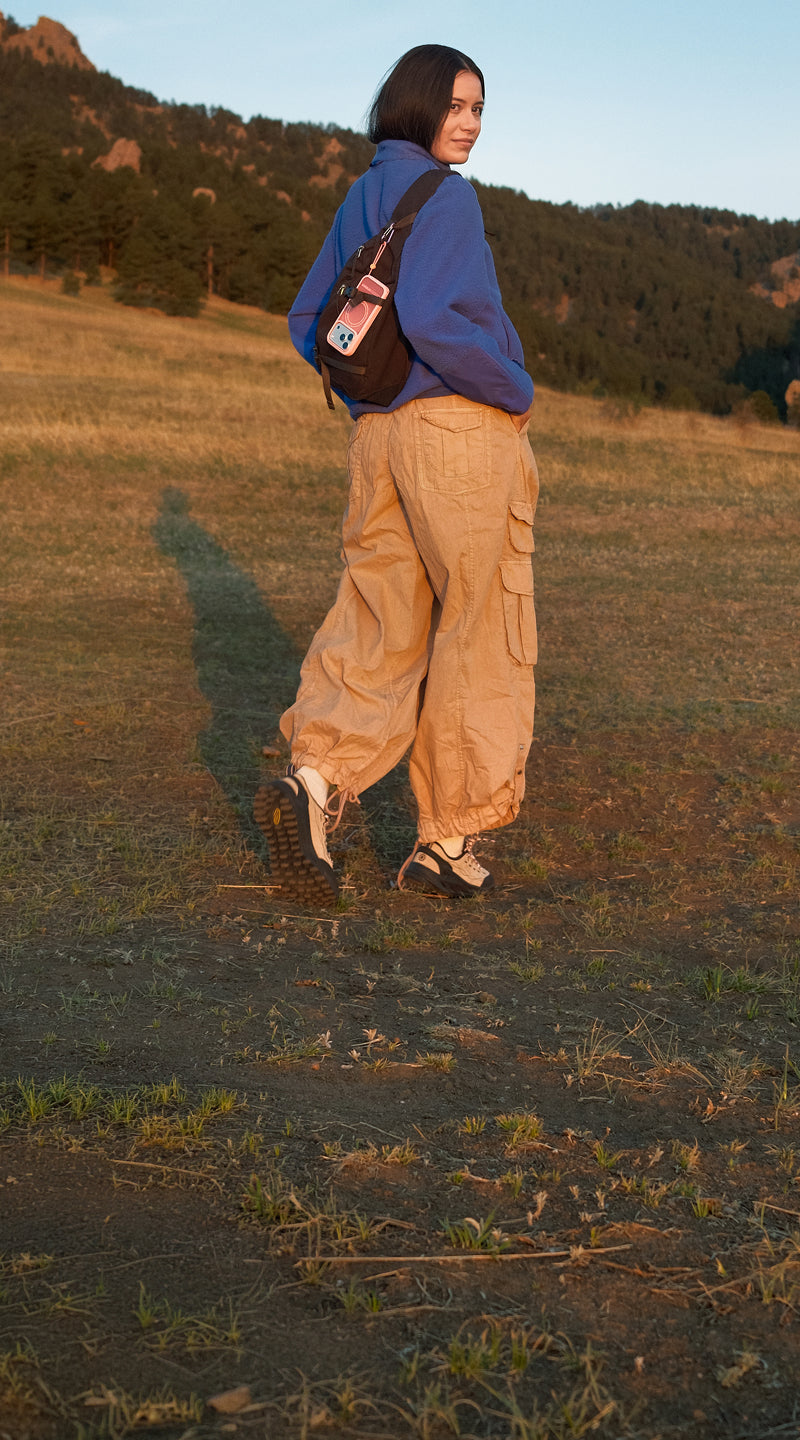 A woman stands in a grassy field with mountains in the background, wearing a blue hoodie, beige cargo pants, and a brown shoulder bag with a Sole series case attached.
