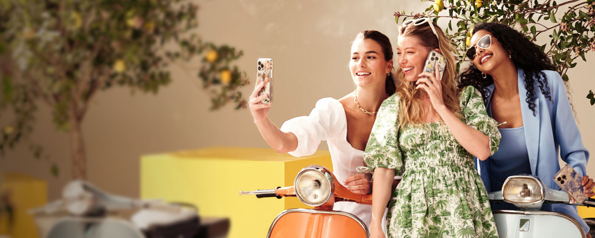 Three women taking a selfie on a scooter with a tree and yellow bench in the background