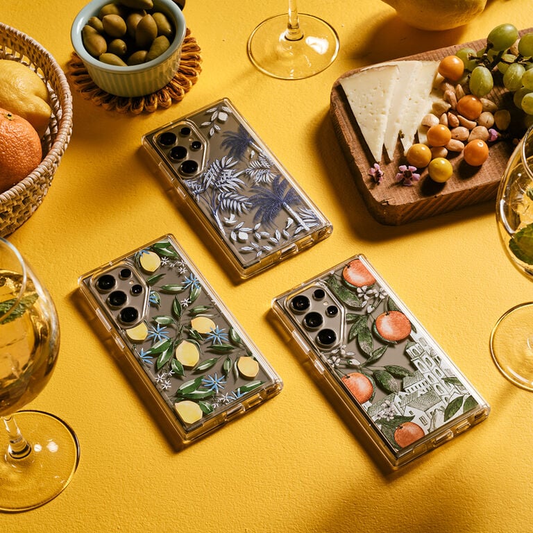 Three phone cases with nature-themed designs on a yellow surface with fruit and wine glasses.