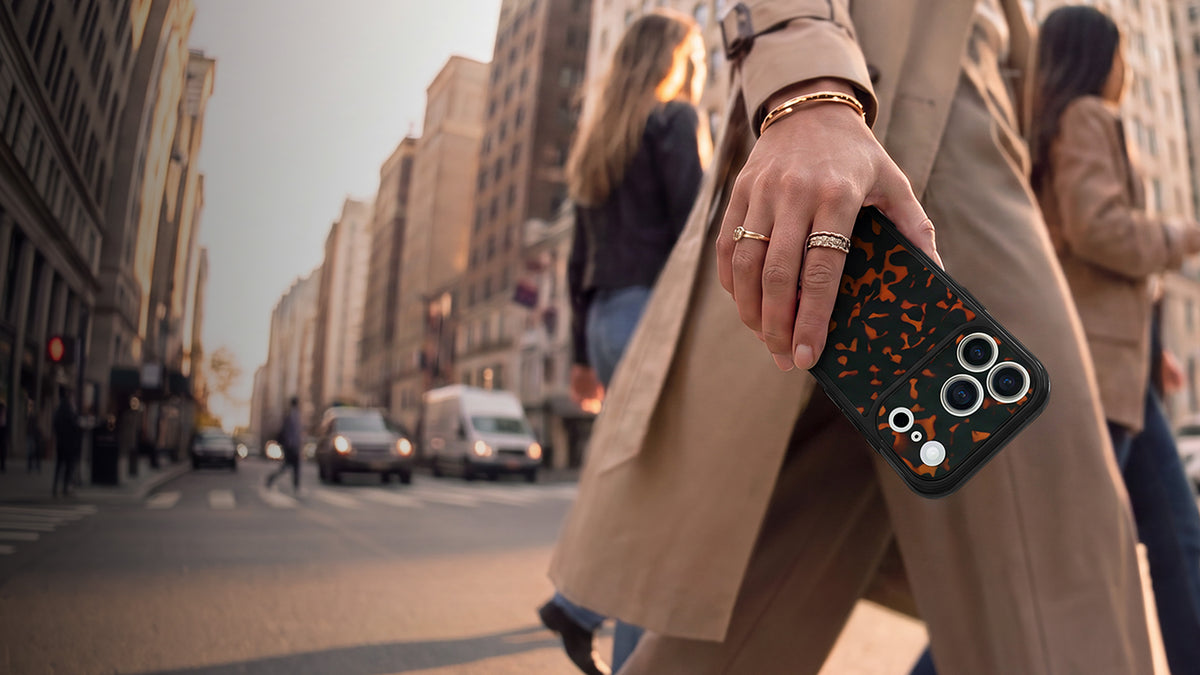 A close-up of a hand holding a black smartphone case with a tortoise print pattern, worn by a person in a beige trench coat on a city street.