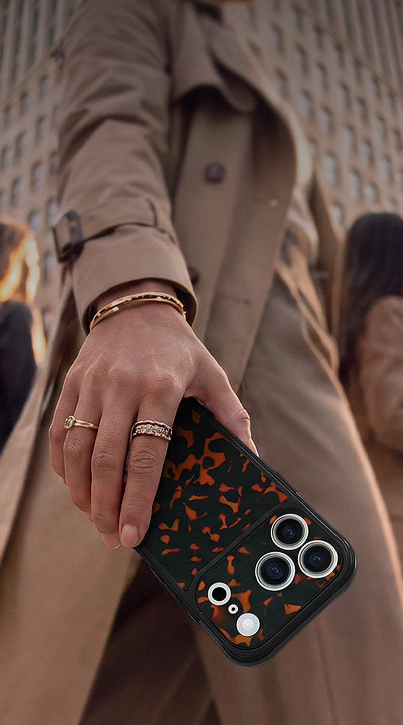A close-up of a hand holding a black smartphone case with a tortoise print pattern, worn by a person in a beige trench coat on a city street.