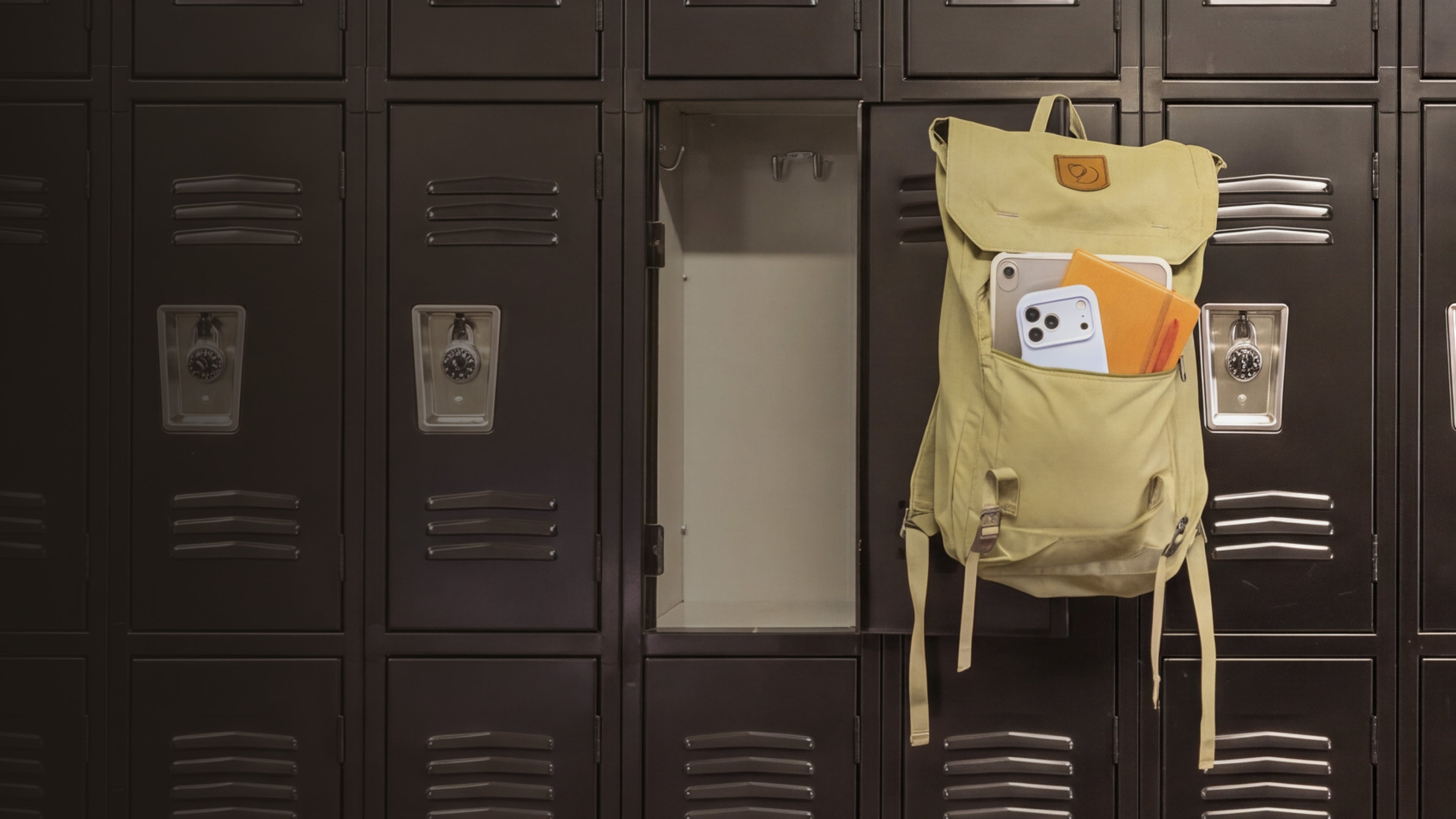 A light beige backpack hanging on a school locker, with a white smartphone and orange folder tucked into its front pockets, set against a row of dark metal lockers.