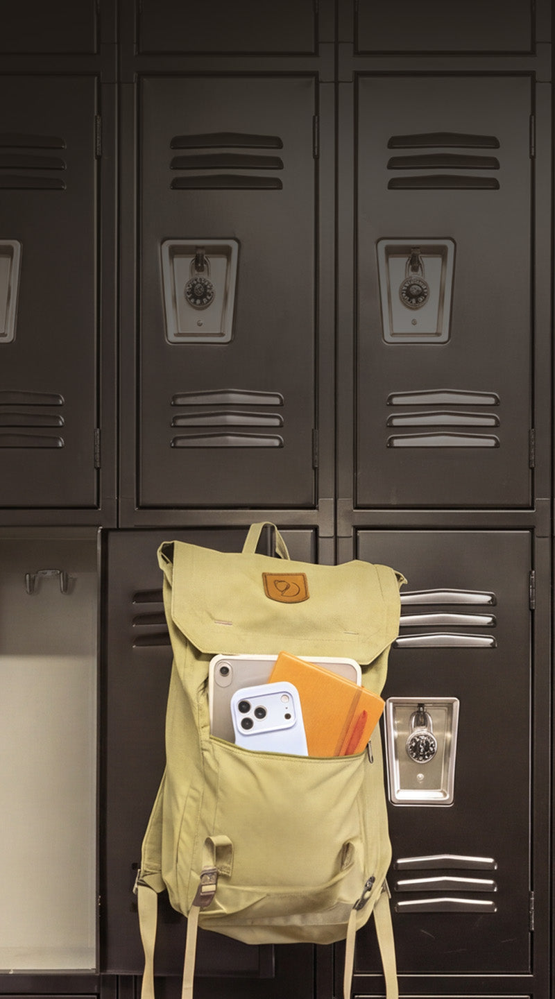A light yellow backpack with a front pocket holding a white smartphone and an orange notebook, hanging in front of dark gray metal lockers.