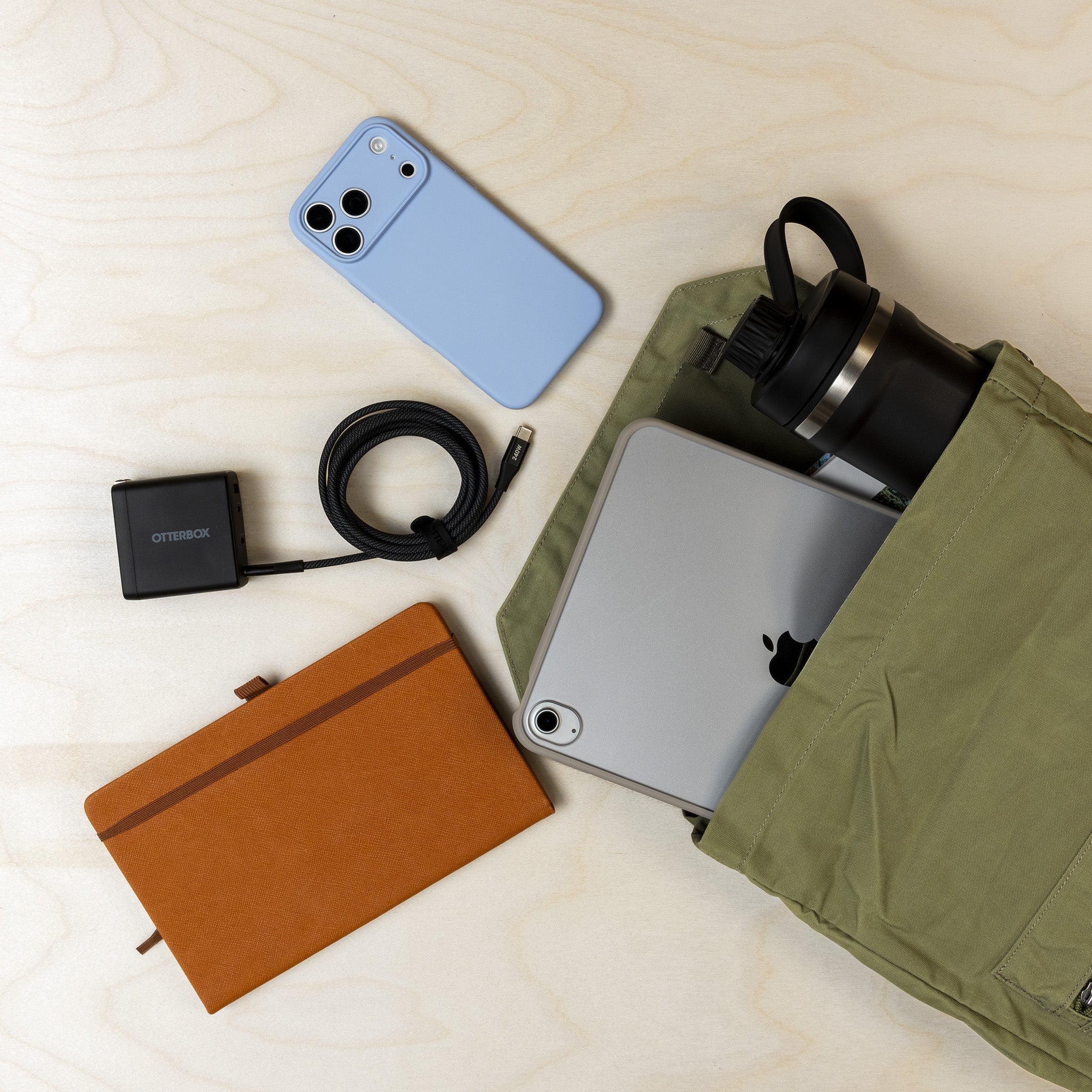 An olive green backpack laid flat on a light wood surface, shown partially open with a black insulated bottle, a silver tablet, a light blue phone case, a black power adapter, and a brown notebook arranged around it.