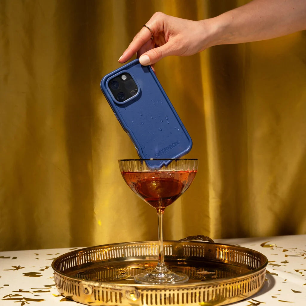 Hand holding a blue phone case above a cocktail on a gold tray with a yellow curtain background
