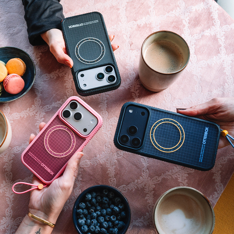 Three phone cases in black, pink, and blue held by hands on a table with food and drinks.