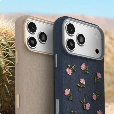 Two smartphone cases shown upright outdoors with a blurred desert landscape and cactus in the background. The left case is a light warm beige textured leather look, and the right case is dark navy with pink floral embroidery and green leaf accents.
