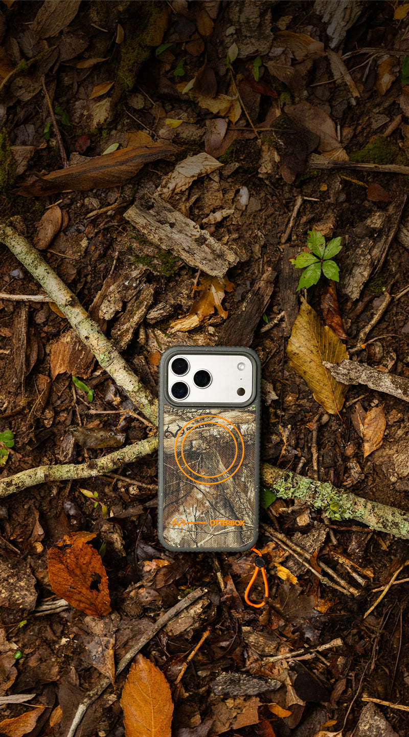 A camouflage phone case with an orange circular logo and text, shown from above on a forest floor covered in leaves, twigs, and soil.
