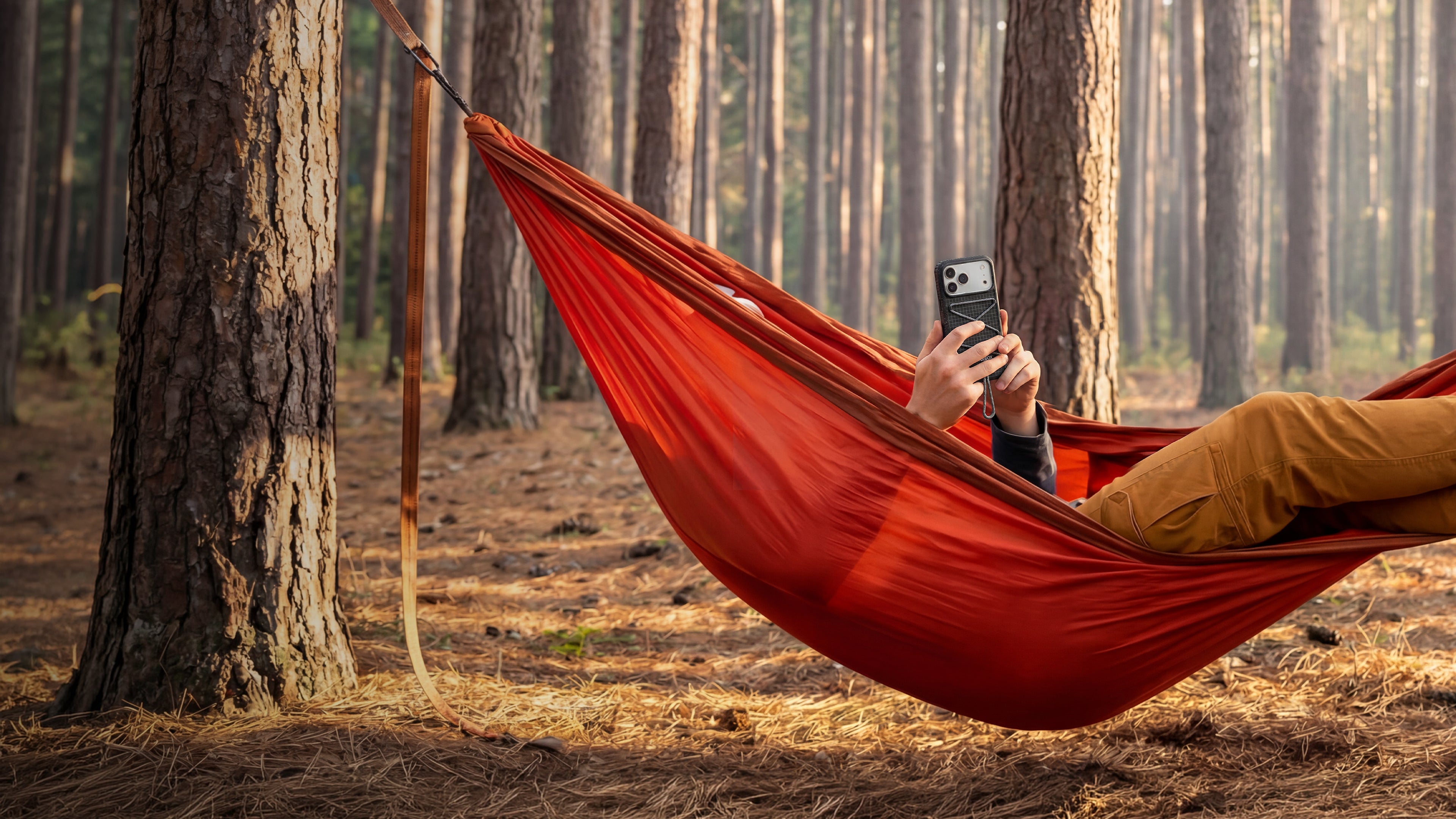 Person relaxing in a red hammock in a forest, using a smartphone.