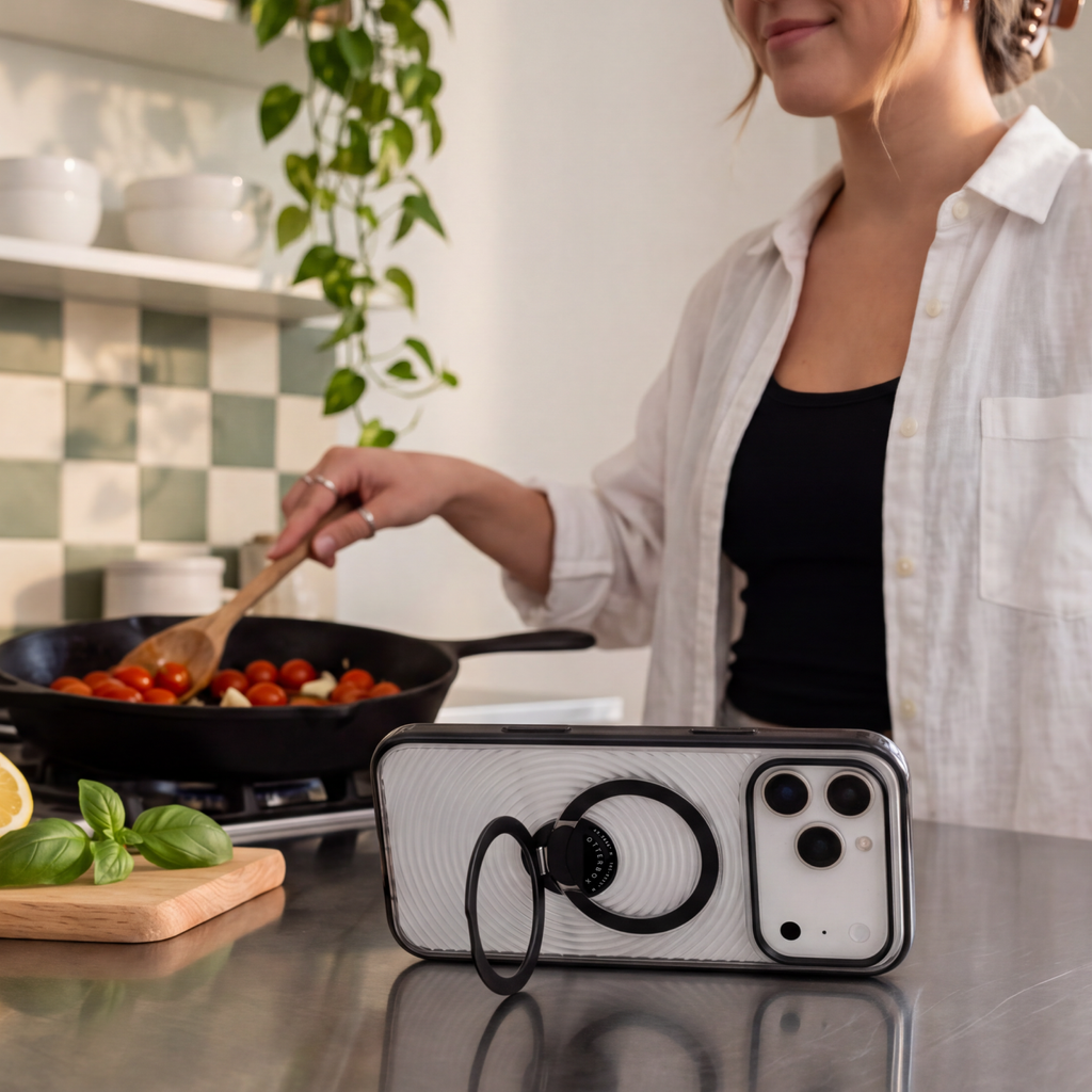 OtterBox Symmetry Series Stand phone case resting on a kitchen counter with its built-in ring stand deployed, while a person cooks in the background.
