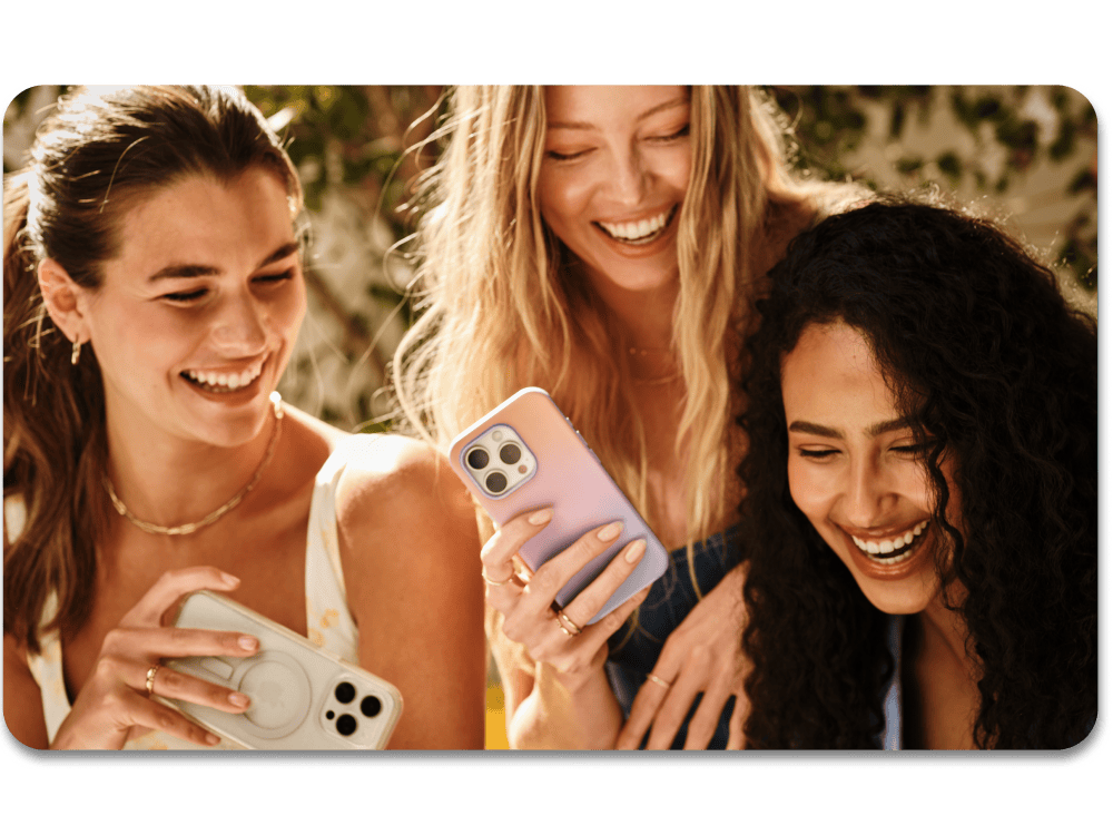Three women laughing together, looking at a phone screen outdoors.